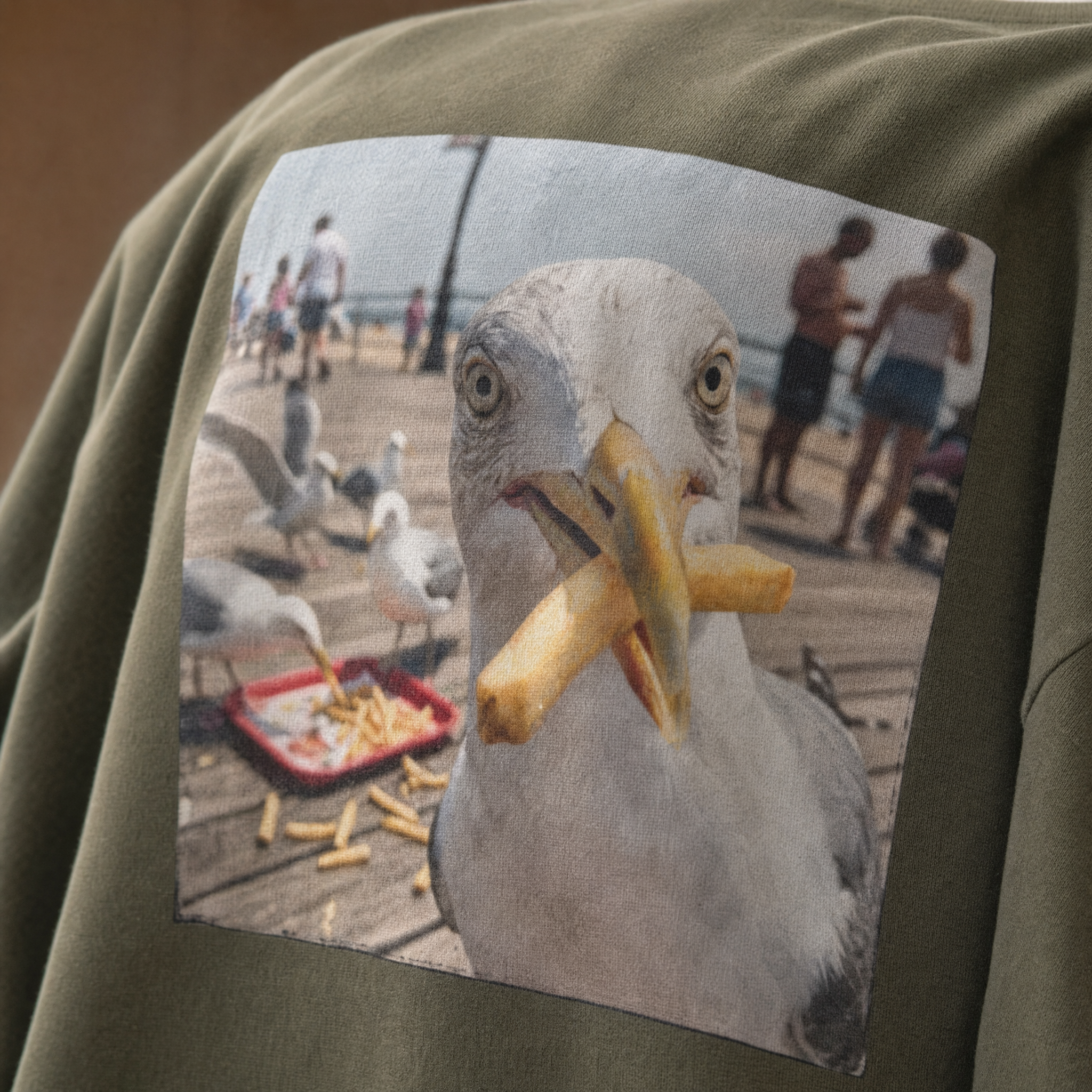 Olive green t-shirt printed with a close-up seagull stealing a chip on a seaside pier